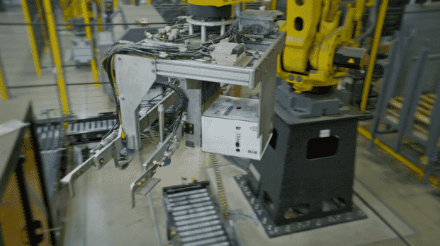 Industrial robotic arm handling materials at a manufacturing plant, with conveyor belts and factory equipment in the background