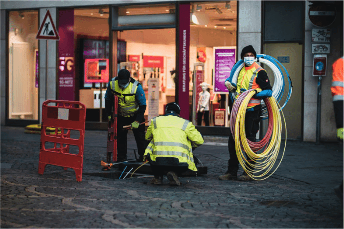 Three workers in high-visibility safety vests and helmets installing fiber optic cables on a city street, highlighting the importance of fiber optic cables for high-speed, reliable internet and data transmission. This image captures the modern infrastructure upgrades required for efficient network performance and the role of fiber optic cables in improving connectivity for various applications, including audio-visual setups, Ethernet networks, and internet infrastructure
