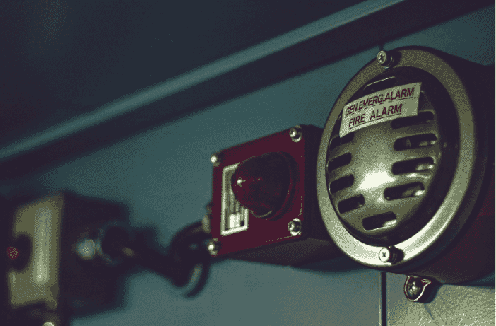 Close-up view of a fire alarm system featuring a circular speaker labeled 'Gen Emerg Alarm Fire Alarm' alongside a red indicator light mounted on a blue industrial panel, symbolizing the importance of reliable safety systems in modern buildings