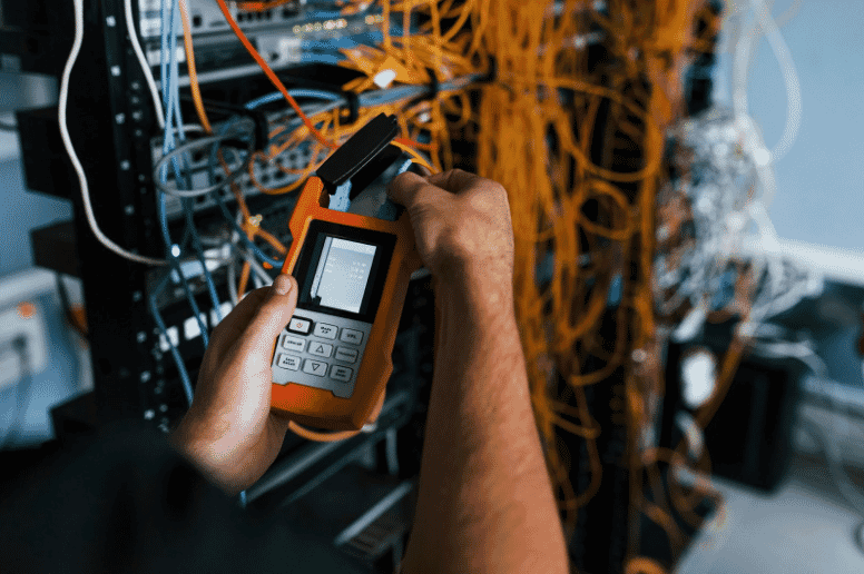Technician using a network cable tester to ensure redundancy, availability, and reliability in a data center setup, surrounded by a network of cables and servers
