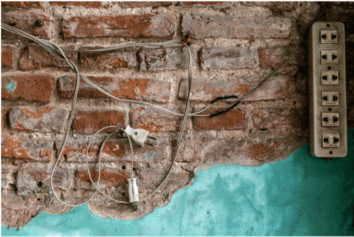 An old brick wall with exposed outdoor electrical wires, showing a mix of outdoor wire and cable installations. The wires include exterior electrical connections and an electric cable for outside use, some hanging loosely, suitable as basic wire for outside applications. The condition emphasizes the need for proper installation of outdoor electrical wire.