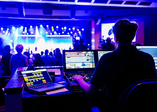 Live event production technician managing audio, video, and lighting systems from a control booth during a stage performance.