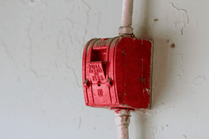 Red manual fire alarm pull station connected to a 2-hour fire rated cable ensuring operational integrity in emergency situations, mounted on a wall with visible signs of wear and age