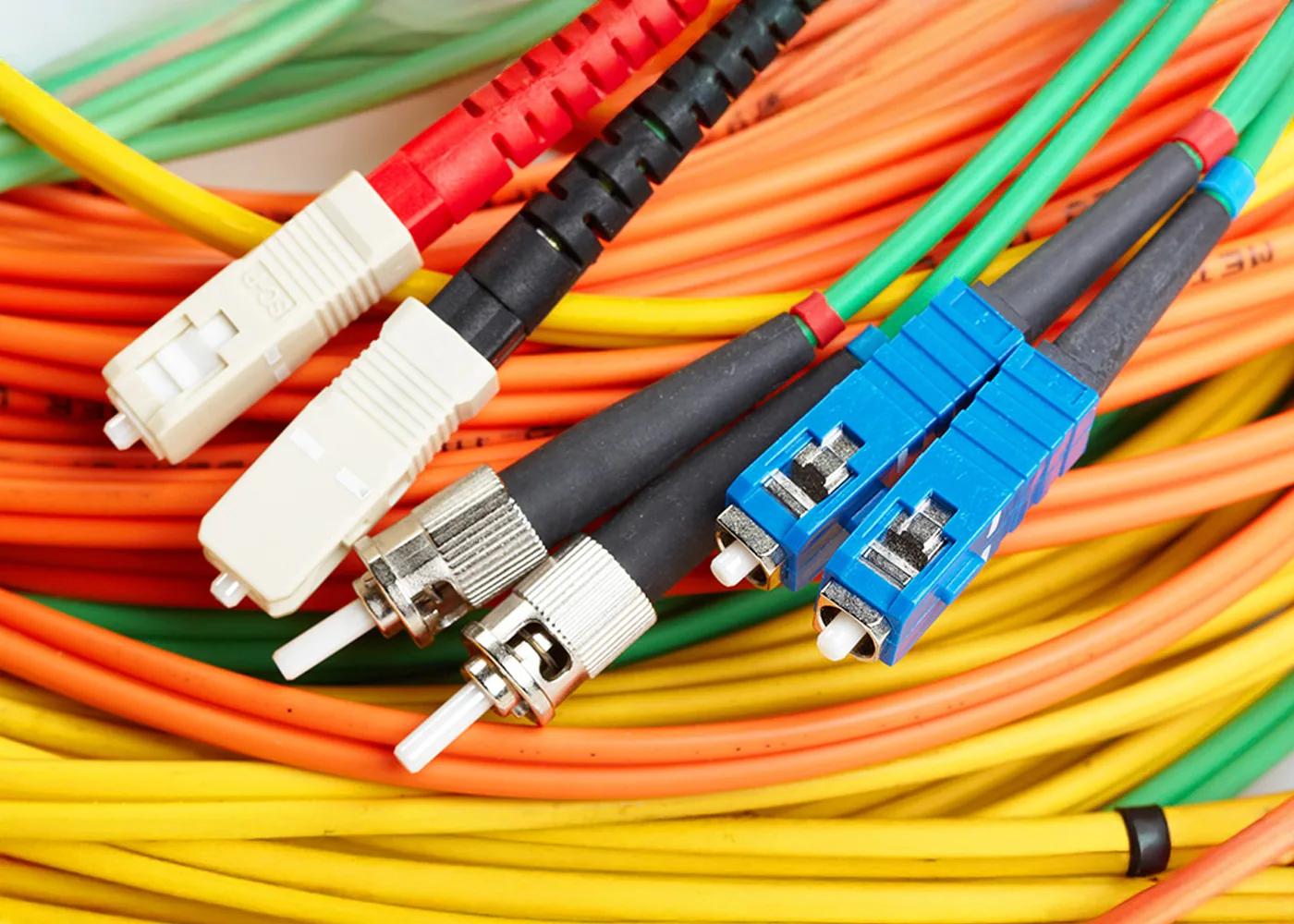 Close-up of colorful fiber optic cables (orange, yellow, and green) with multiple connector types, including blue, white, and metal-tipped ends, bundled together in a network wiring setup.