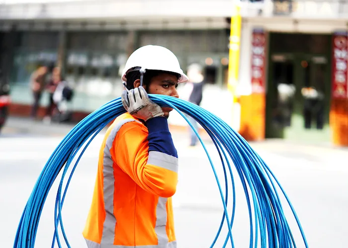Construction worker carrying blue cables wearing safety gear on a city street.